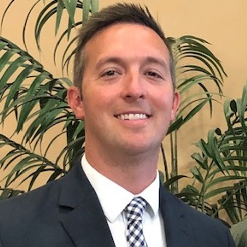 A man with short brown hair smiling, wearing a dark blue suit, white shirt, and blue checkered tie against a leafy plant.