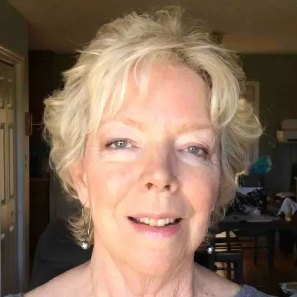A person with short, blonde, wavy hair smiles gently at the camera, indoors with a softly blurred background.