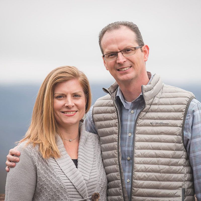 A smiling couple poses for a portrait outdoors, wearing sweaters and vests against a blurred background.