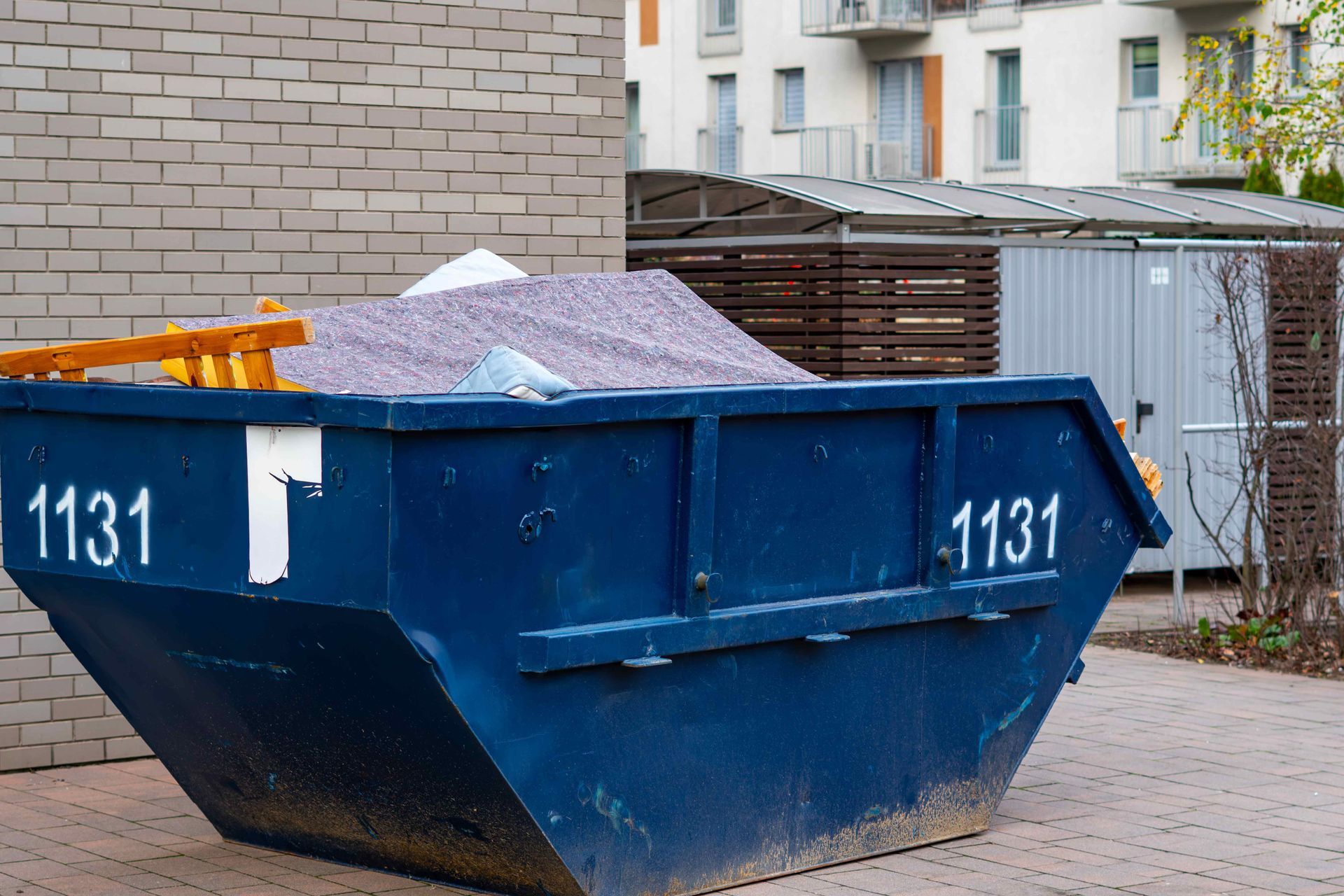 Blue dumpster filled with gravel, numbered 1131, beside a building and fence Blue dumpster filled with gravel, numbered 1131, beside a building and fence