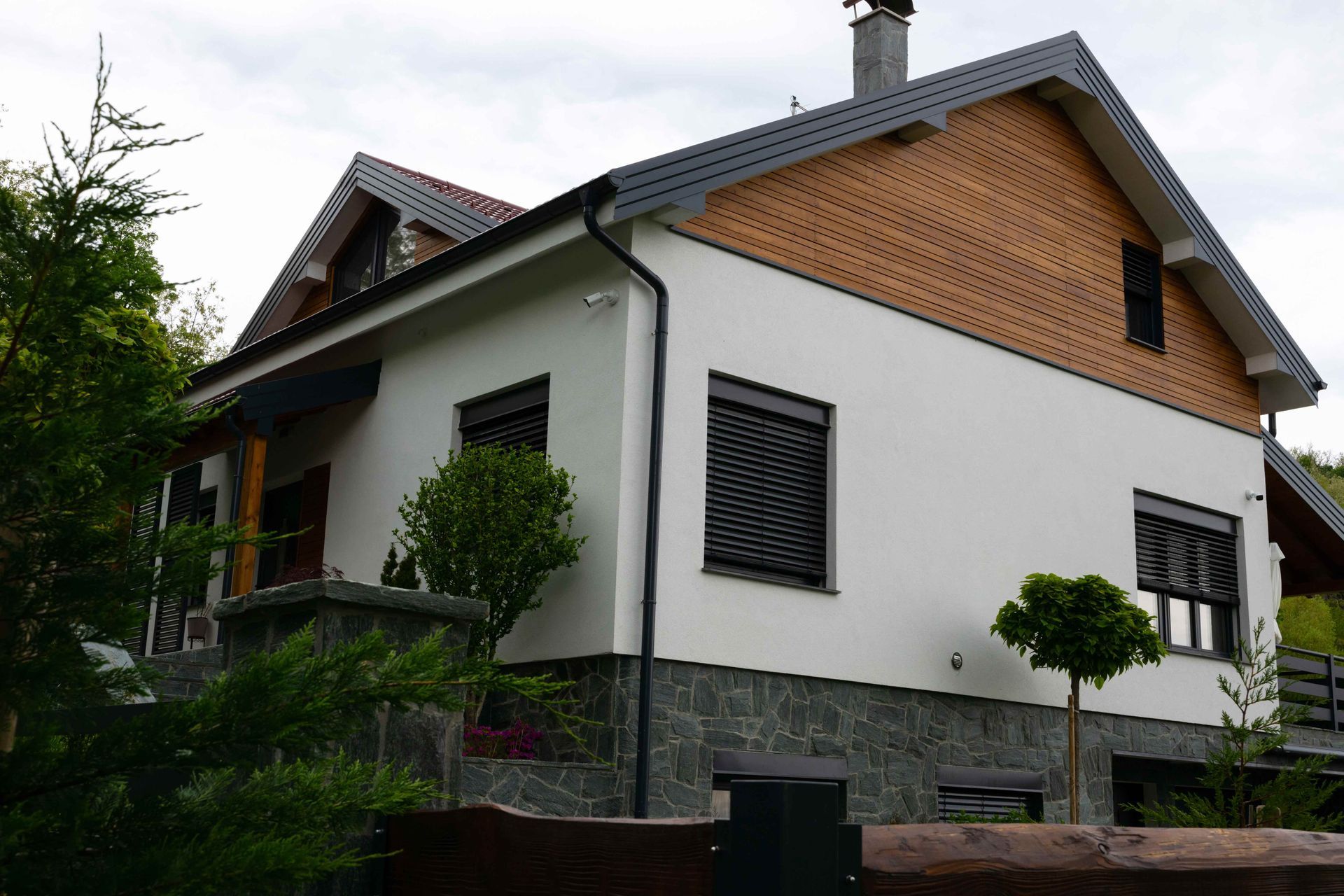 Two-story house with white walls, brick gable, dark roof, and stone base among trees Two-story house with white walls, brick gable, dark roof, and stone base among trees