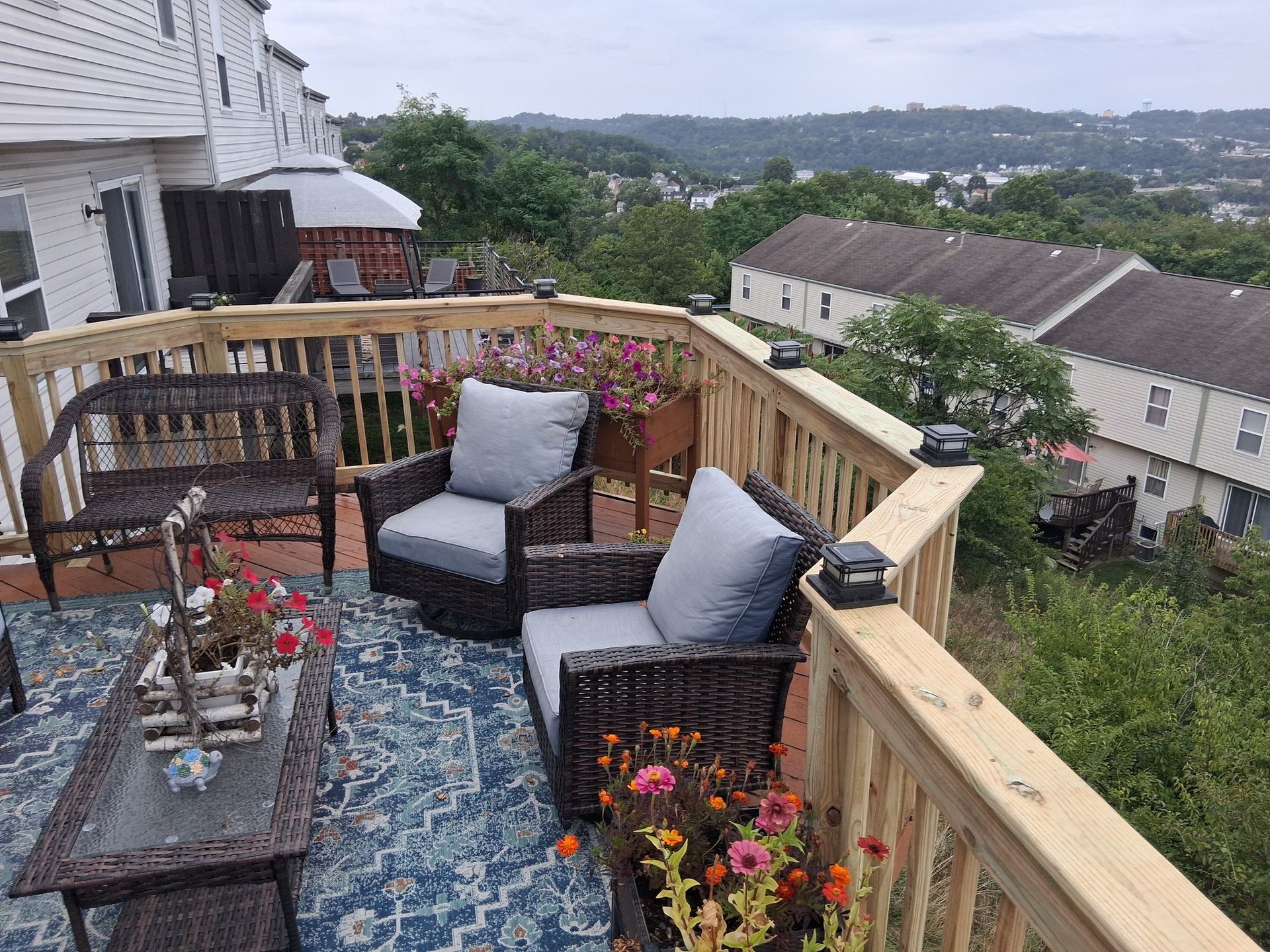 Balcony patio with wicker chairs, blue rug, potted flowers, and a scenic hillside neighborhood view