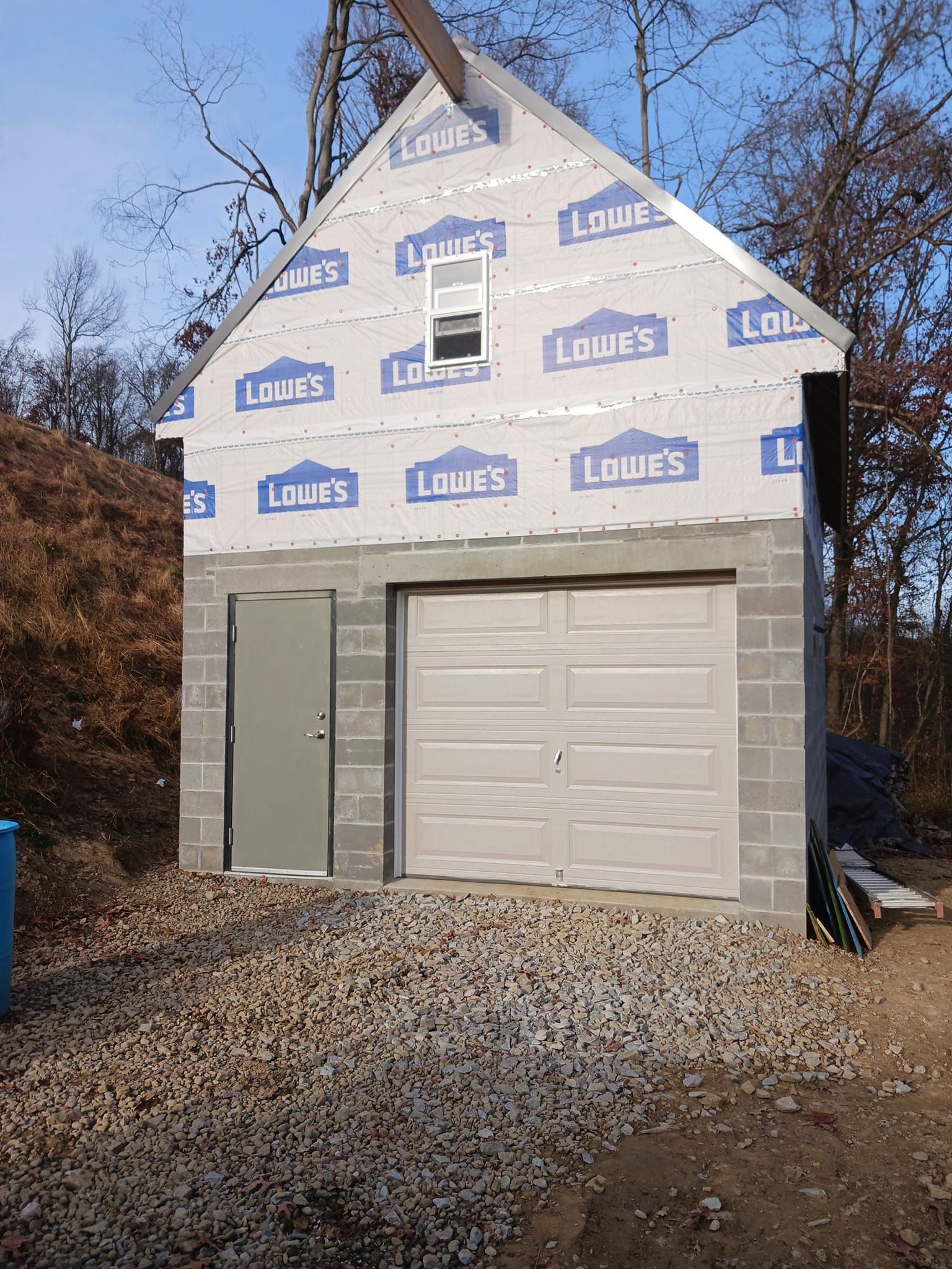 House under construction with white Tyvek wrap, gray garage door, and gravel driveway.