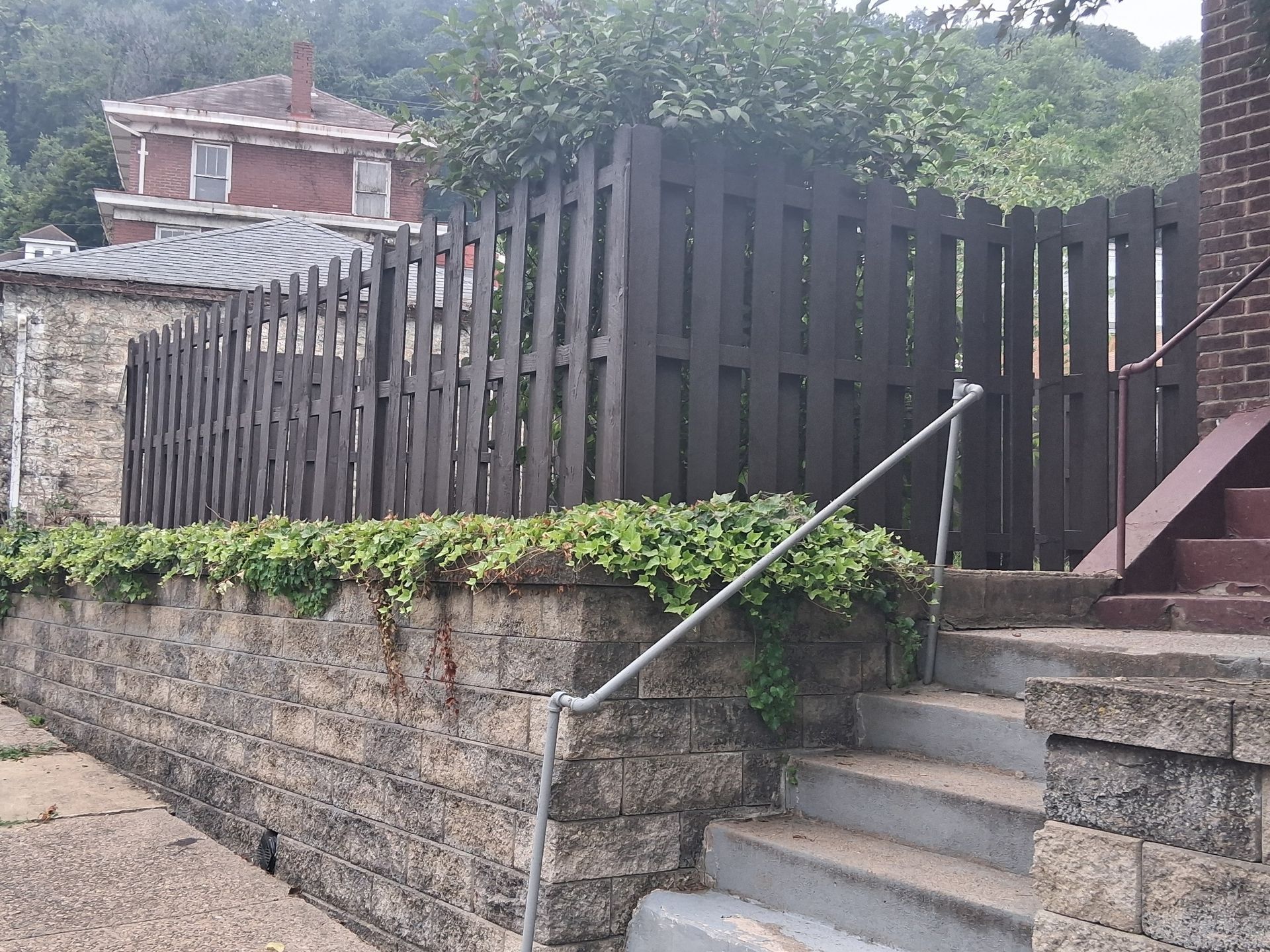 Stone steps and retaining wall with metal handrail beside a dark wooden fence and brick house.