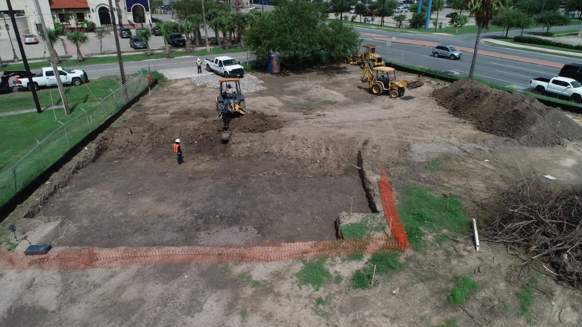 An aerial view of a construction site with heavy machinery, piles of dirt, and a worker standing in a cleared area.