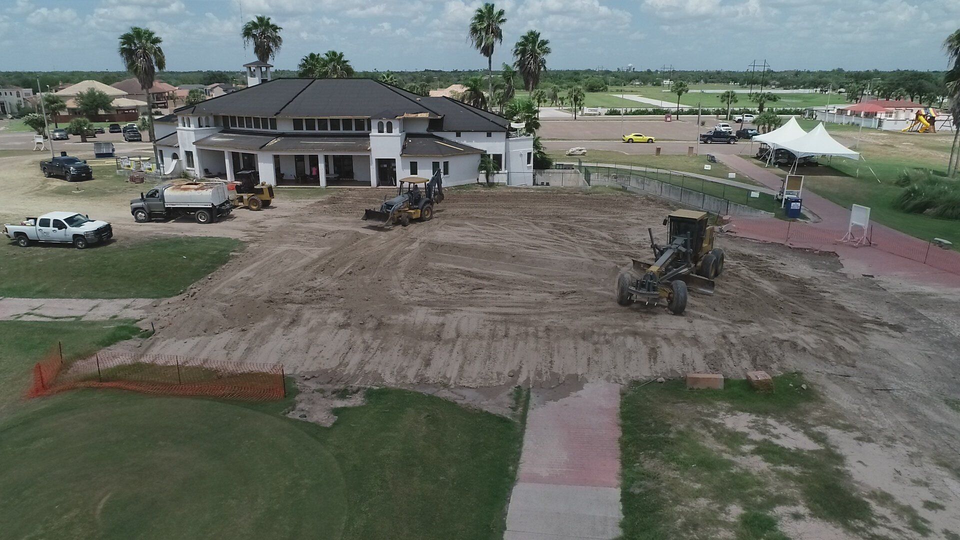 Construction equipment levels a large, muddy dirt patch in front of a white building with a dark roof on a sunny day.