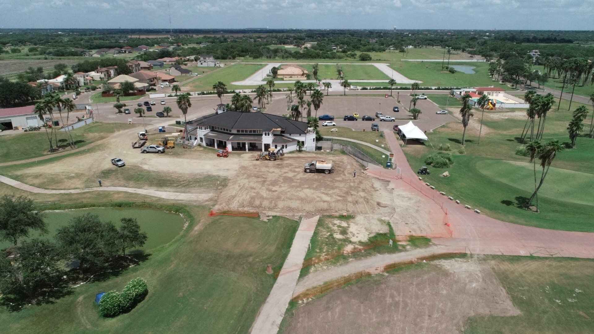 Aerial view of a golf course clubhouse with surrounding parking, palm trees, and an area of disturbed soil under renovation.