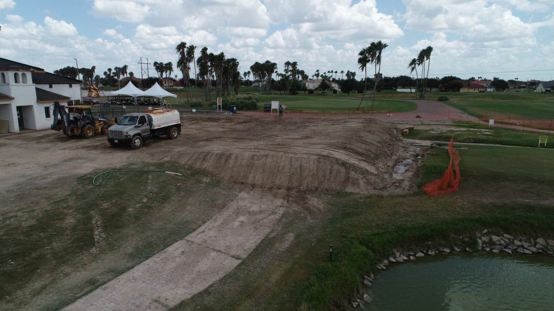 A construction site with dirt mounds, a truck, and heavy machinery near a building and a small pond under a cloudy sky.