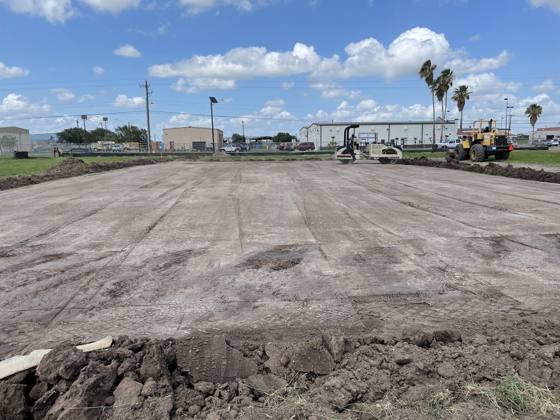 Construction site with a dirt lot prepared for paving, featuring a road roller and tractor under a bright, cloudy sky.