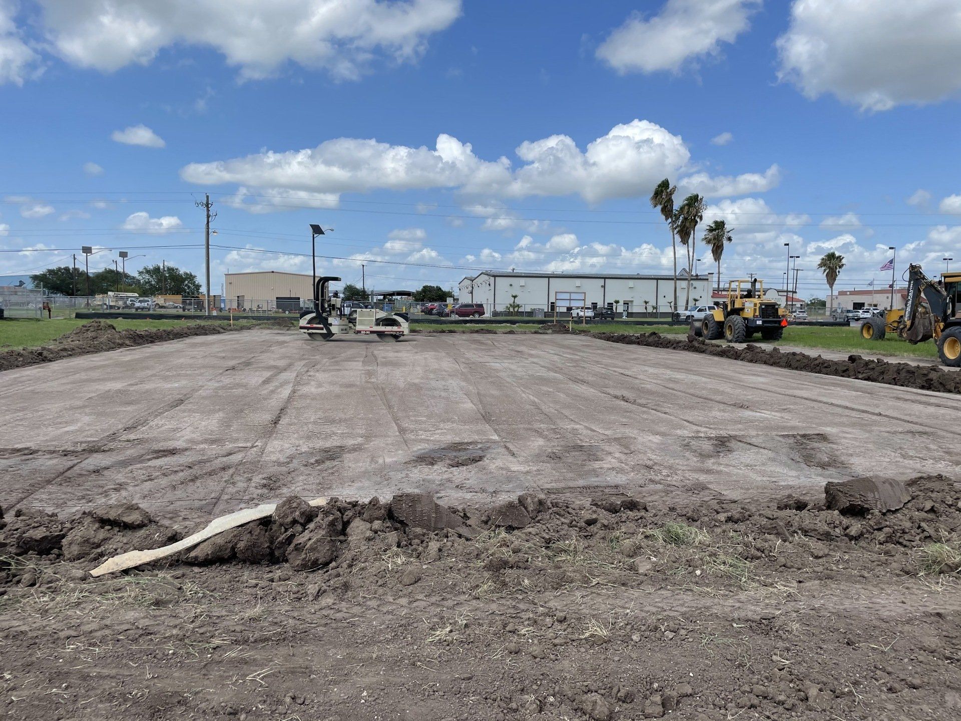 A construction site with a freshly graded dirt lot, heavy machinery, and palm trees under a partly cloudy sky.