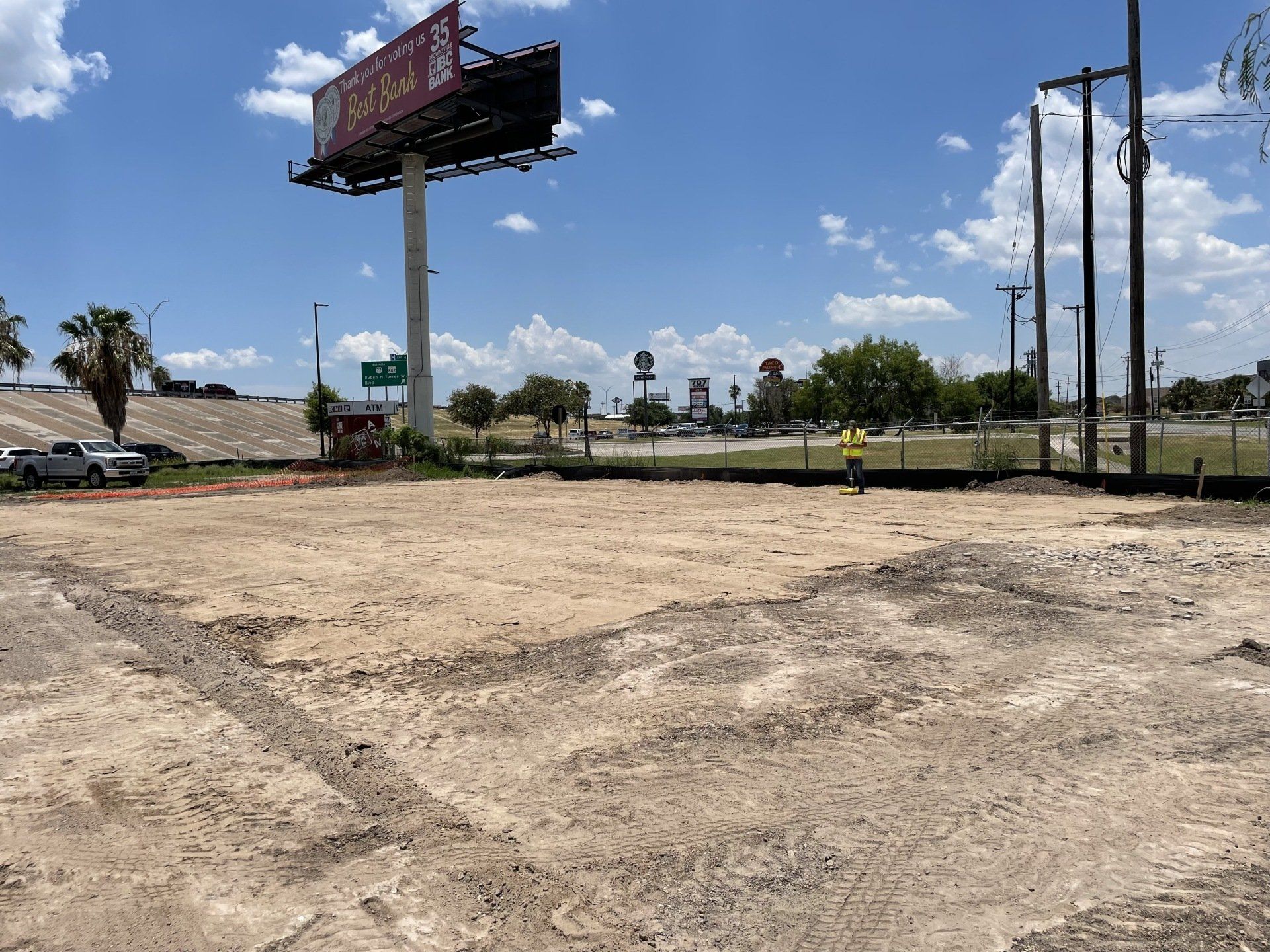 A construction worker stands on a cleared, sandy dirt lot near a billboard and utility poles under a sunny blue sky.