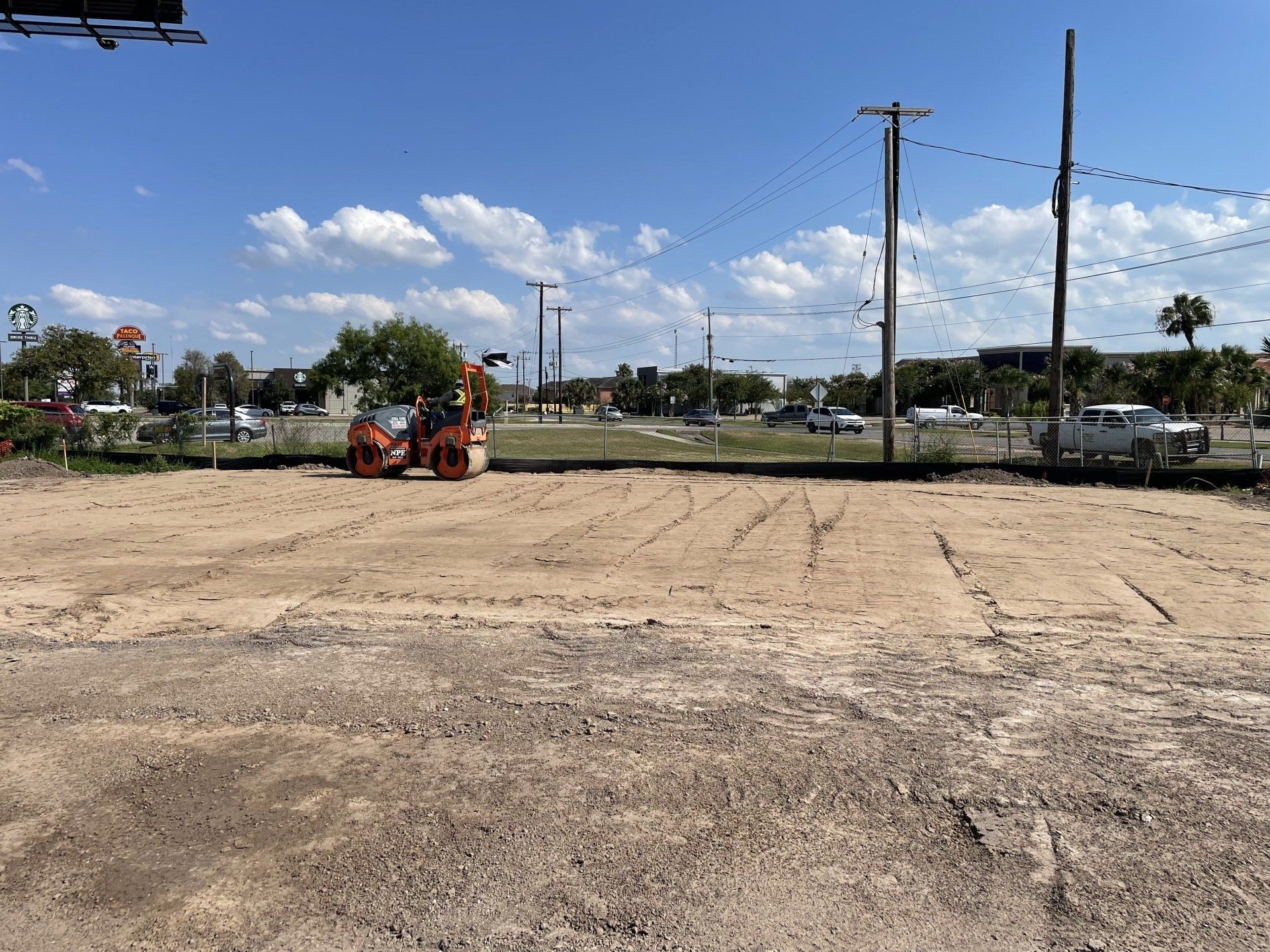An orange construction roller levels a dirt-covered vacant lot under a sunny blue sky with scattered clouds.