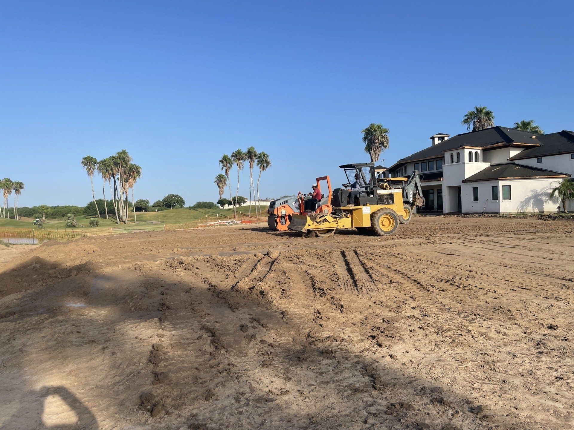 A yellow construction tractor levels dirt on a site near a white house under a clear blue sky.