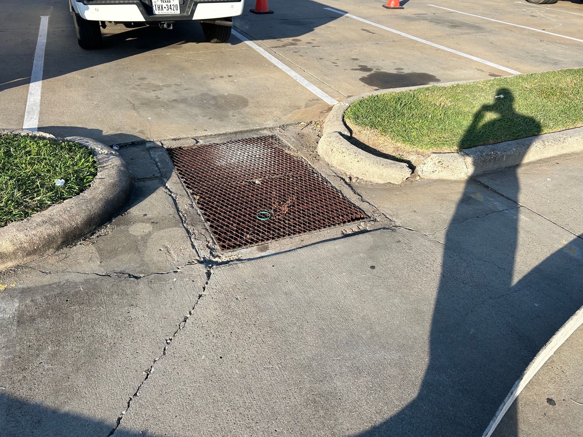 A rectangular metal storm drain grate sits in a concrete parking lot between two patches of grass.