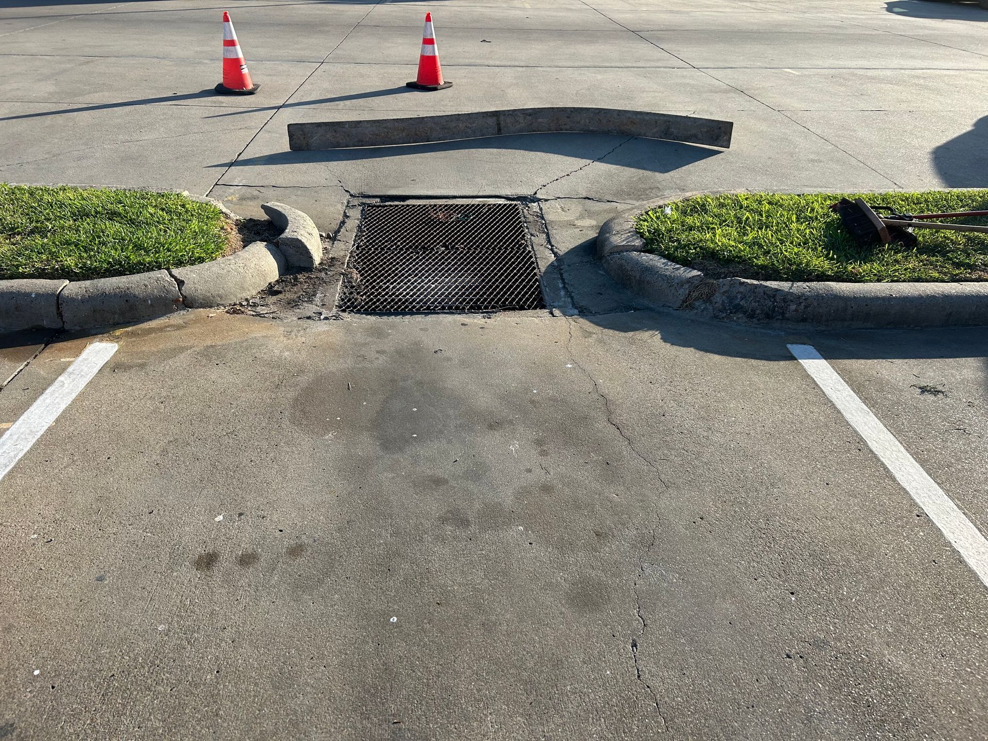 A storm drain grate located in a parking lot between two landscaped curbs, with two traffic cones placed in the distance.