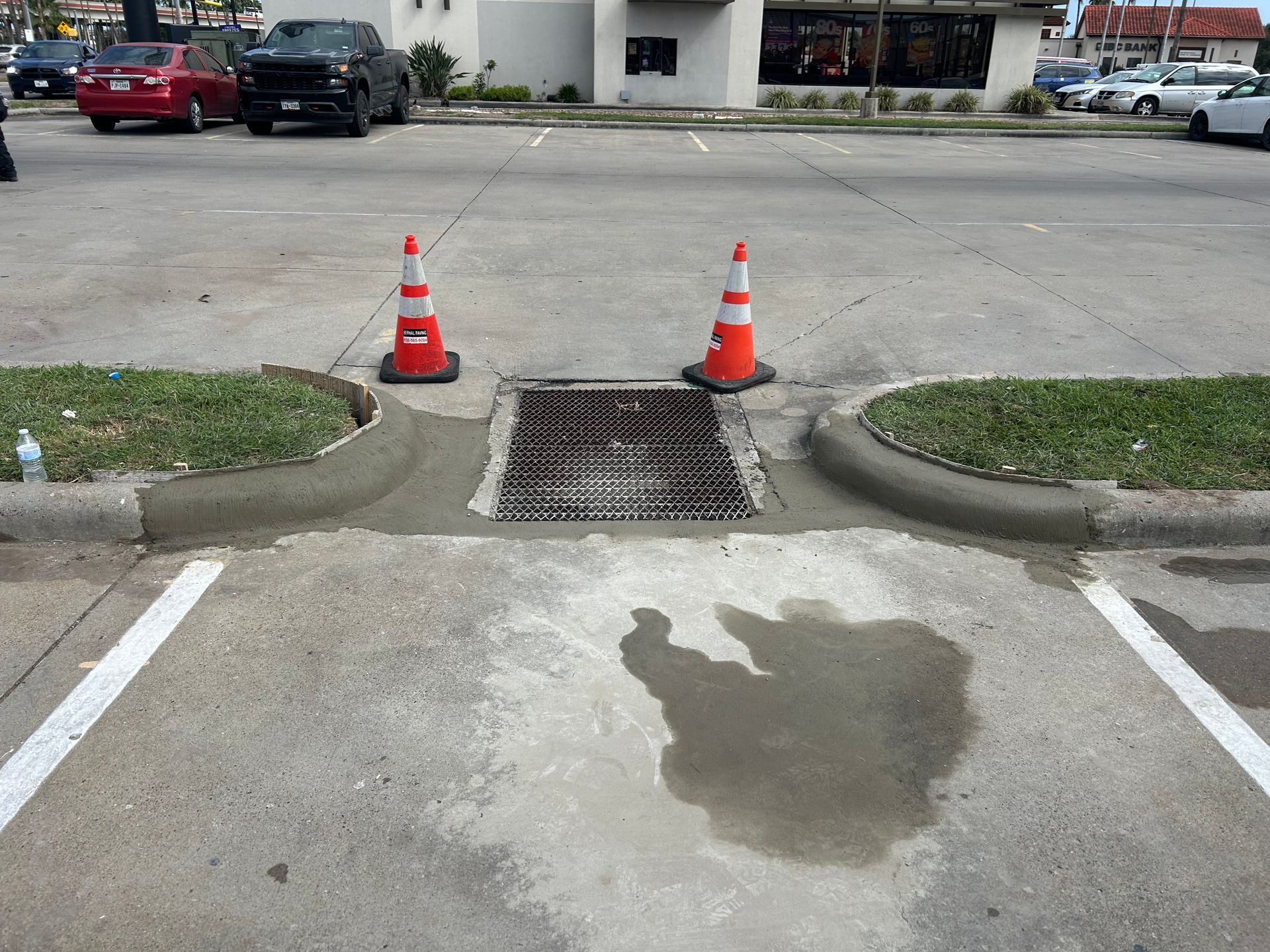 A storm drain grate set between two concrete curbs in a parking lot, marked by two orange and white traffic cones.