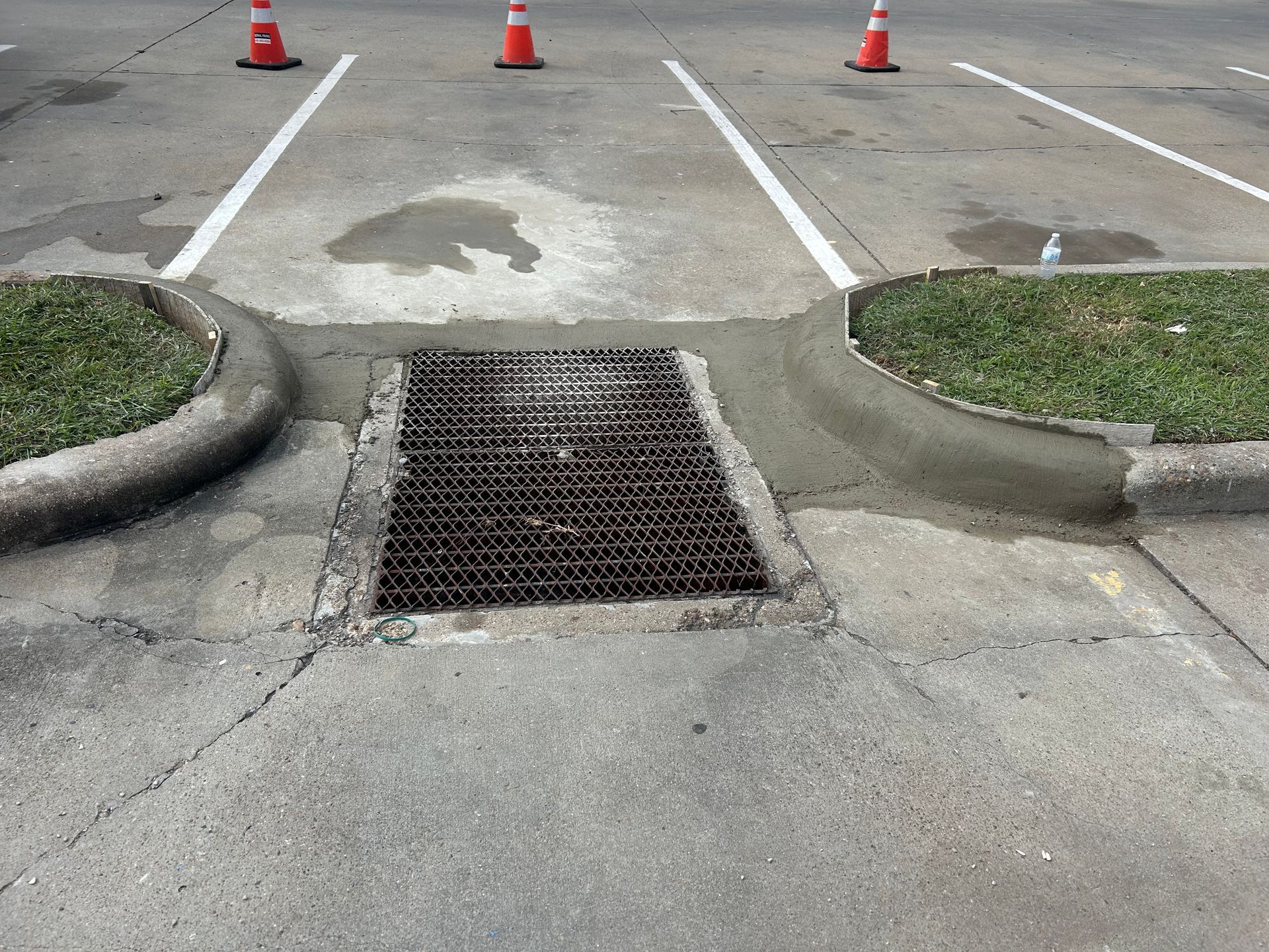A metal storm drain grate located between two concrete curbs in a parking lot, with three orange traffic cones in the back.