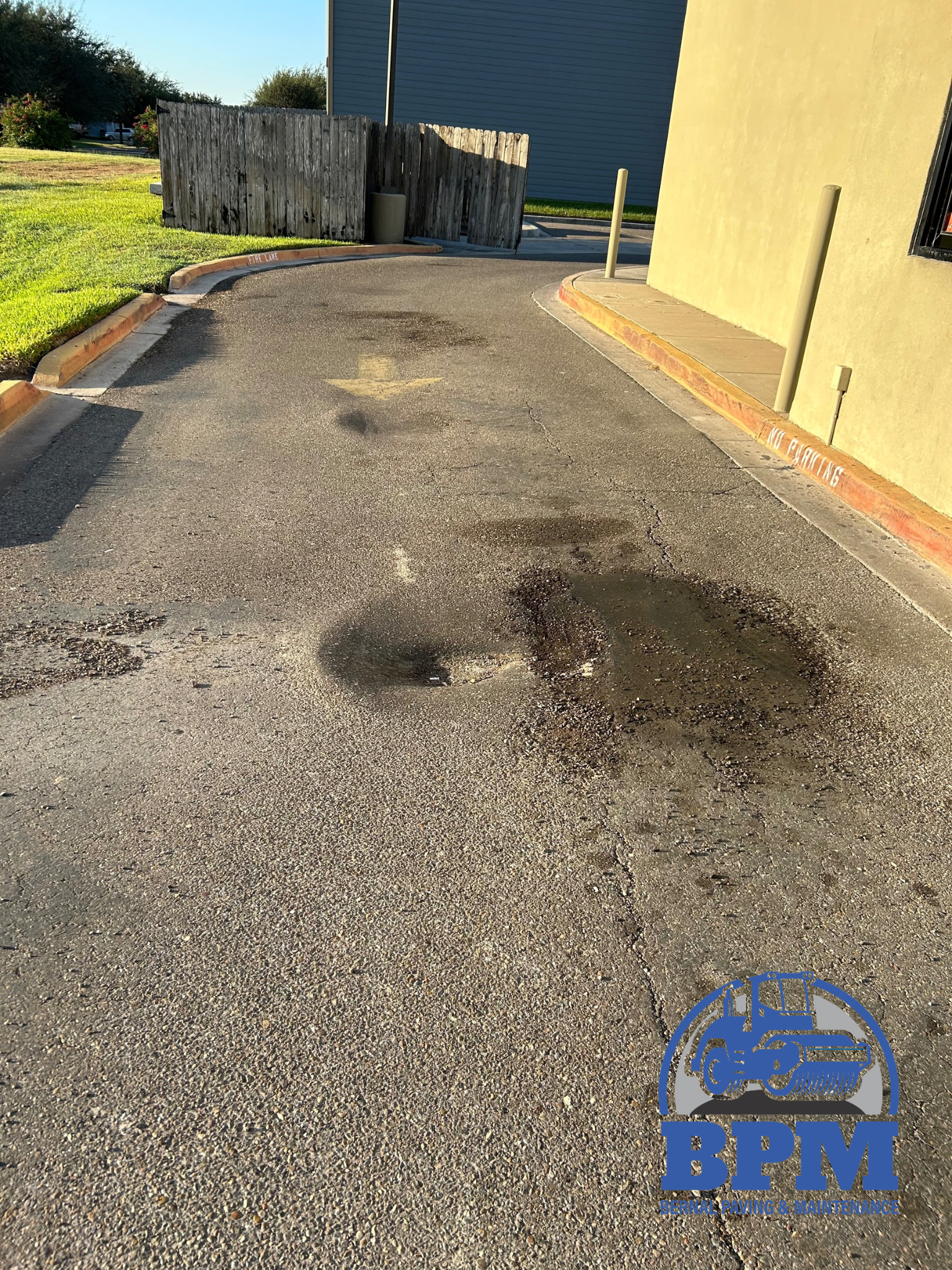 A paved area showing potholes, cracking, and water pooling next to a building and a wooden fence.