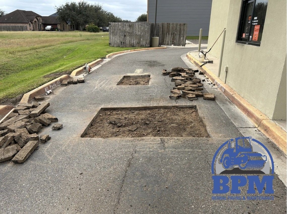 Construction work in progress with two rectangular holes cut into an asphalt driveway alongside a commercial building.