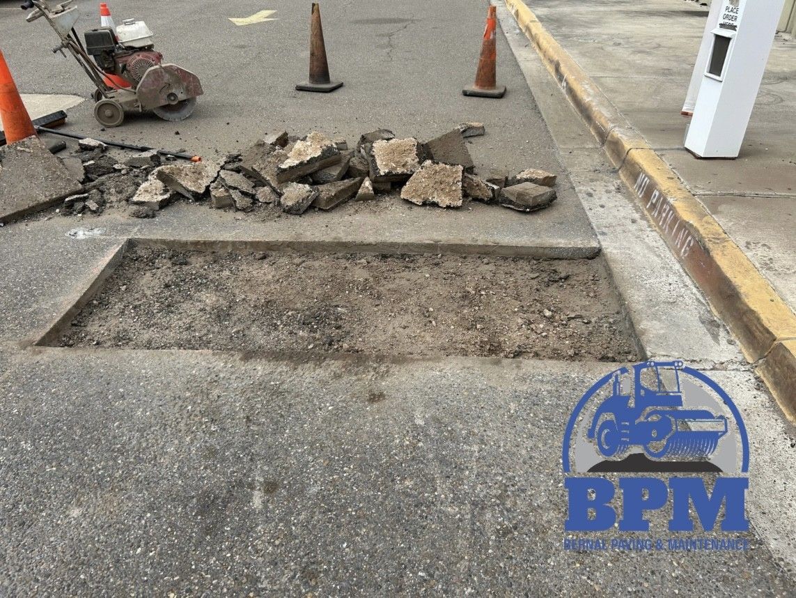 A rectangular hole cut into an asphalt parking lot, surrounded by concrete debris, traffic cones, and construction tools.