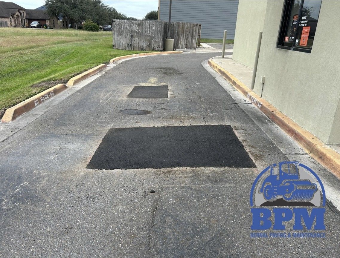 Asphalt driveway area showing two recent, rectangular pavement patches and concrete curbs, with a logo in the corner.