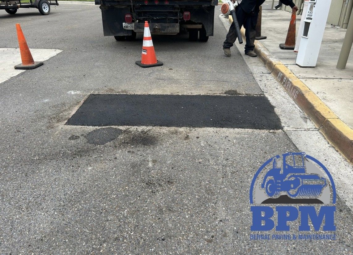 A worker blows debris off fresh asphalt pavement near a truck and orange safety cones.