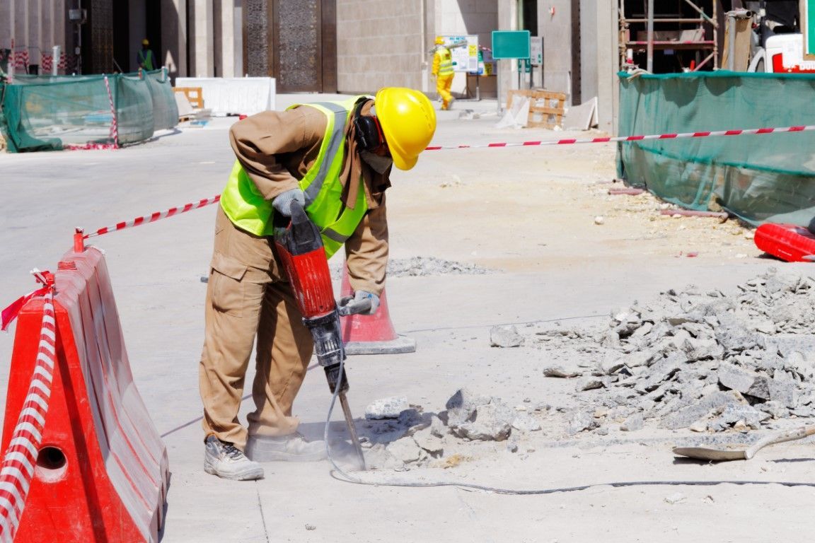 A construction worker in a yellow hard hat and neon vest uses a jackhammer on concrete at a site.