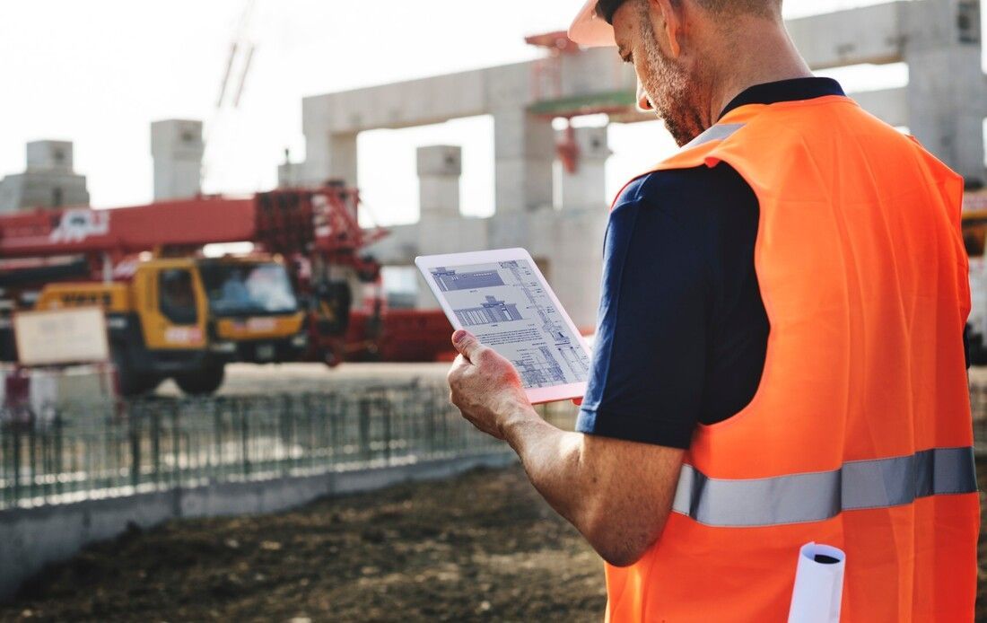A construction worker in an orange vest and helmet reviews blueprints on a tablet at a building site.