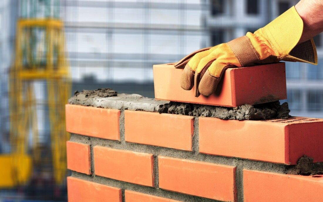 A gloved hand carefully places a red brick onto a partially built wall with fresh mortar.