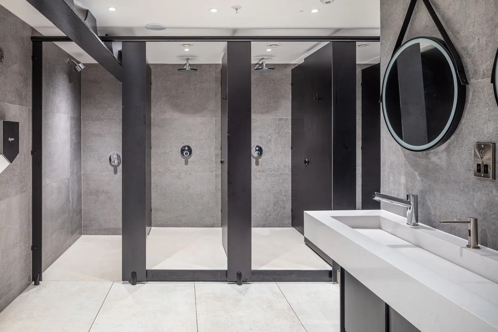 Public shower room with gray concrete walls, black dividers, white sink and mirror.