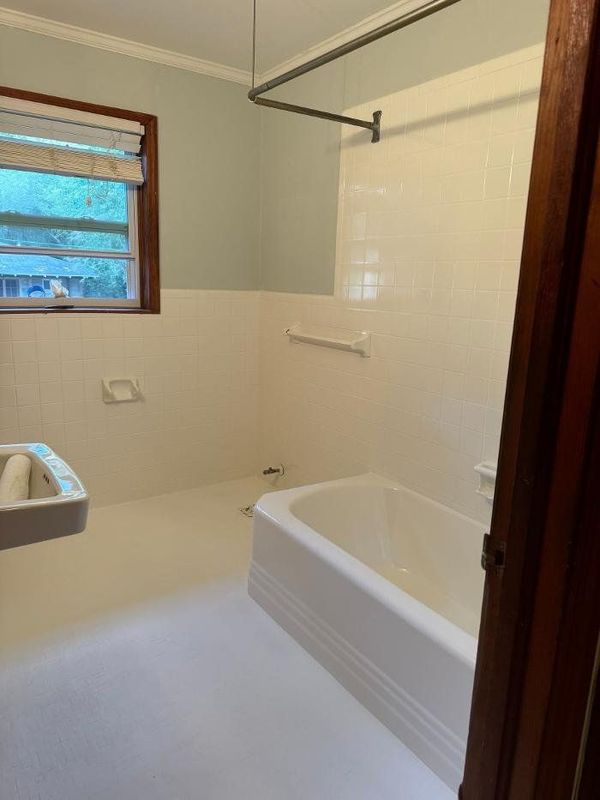 A small bathroom with a white tub, tile, and floor. Window with a shade and a blue wall.