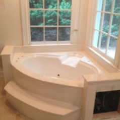 Corner whirlpool bathtub with two steps, set beneath windows in a white bathroom.
