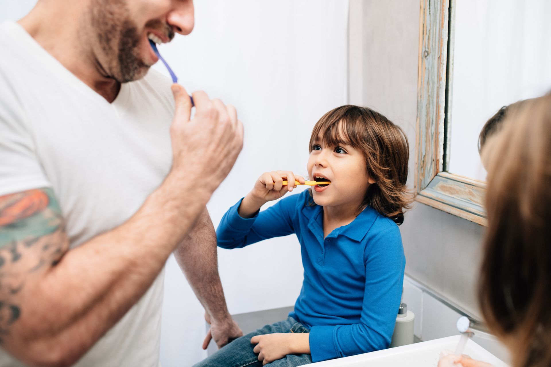Un uomo e un bambino si lavano i denti in un bagno. L'uomo ha dei tatuaggi e sorride. Il bambino è seduto, concentrato.