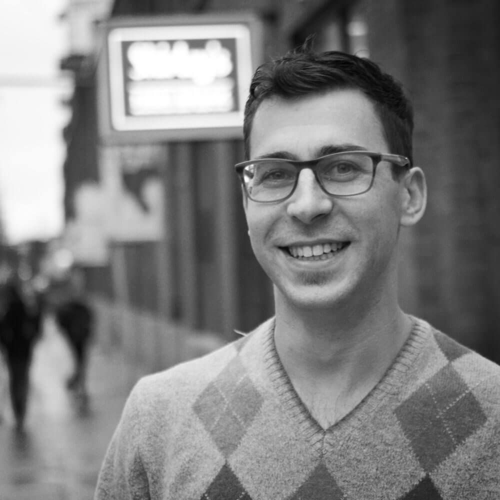A man wearing glasses and a plaid sweater is smiling in a black and white photo.