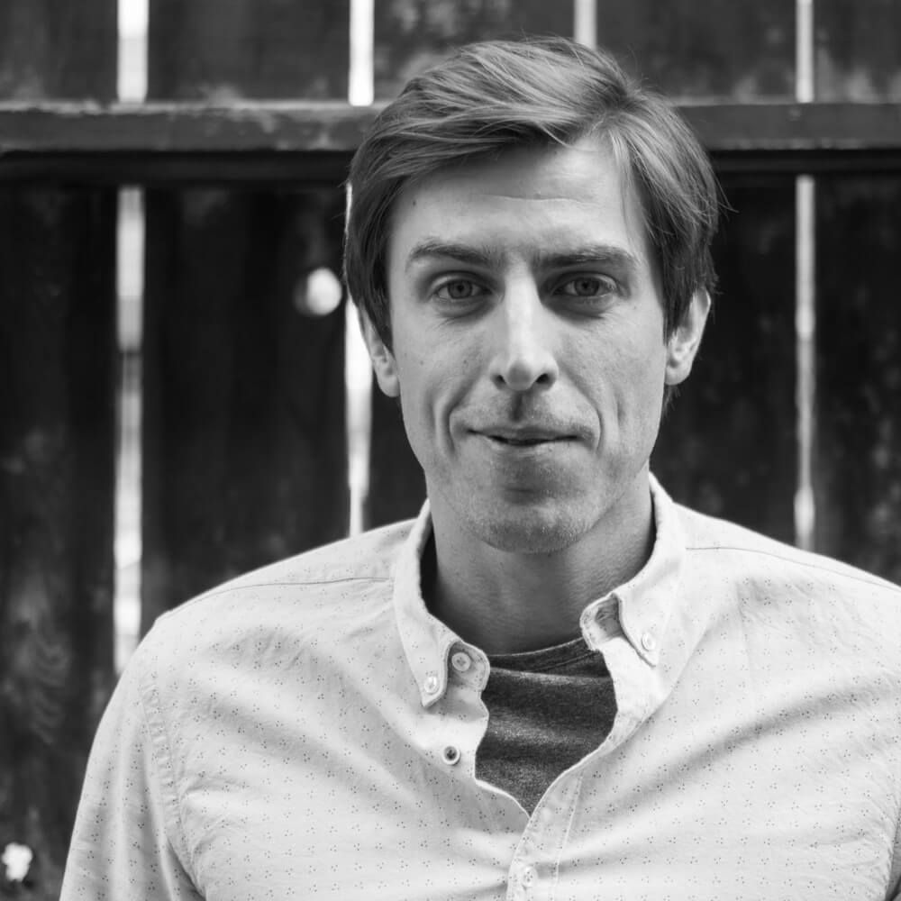 A man in a white shirt is standing in front of a wooden fence.