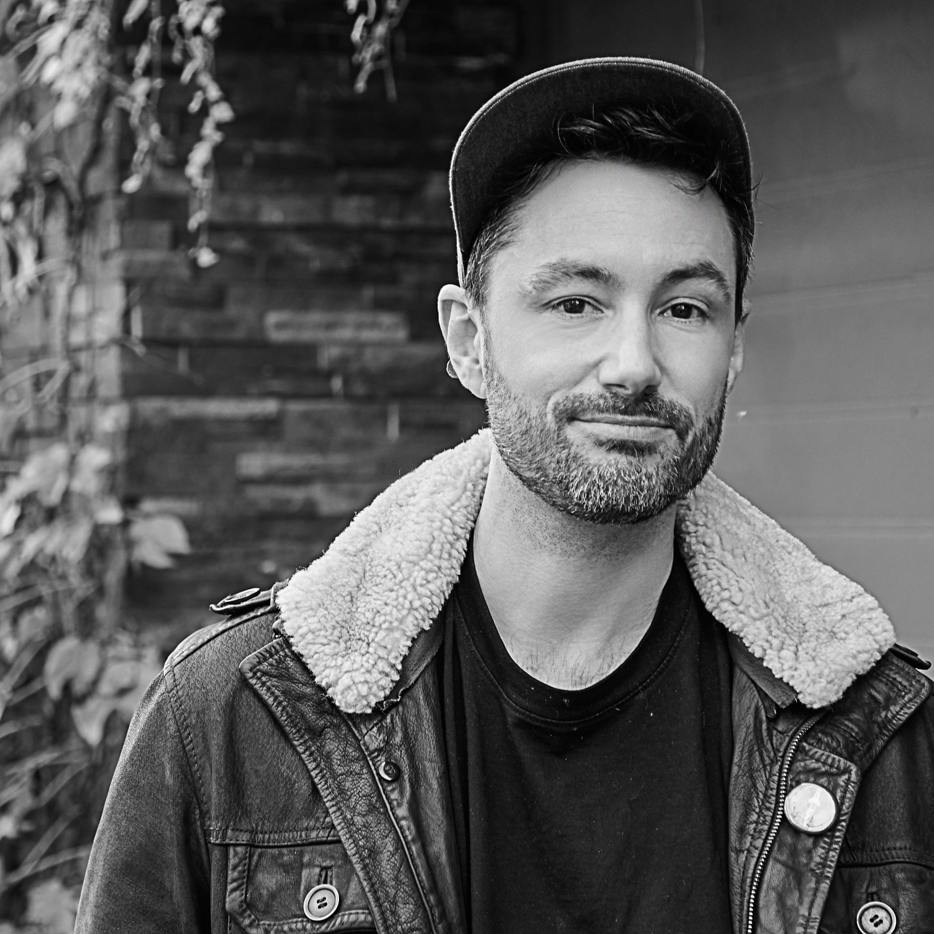 A man with a beard is wearing a hat and a leather jacket.