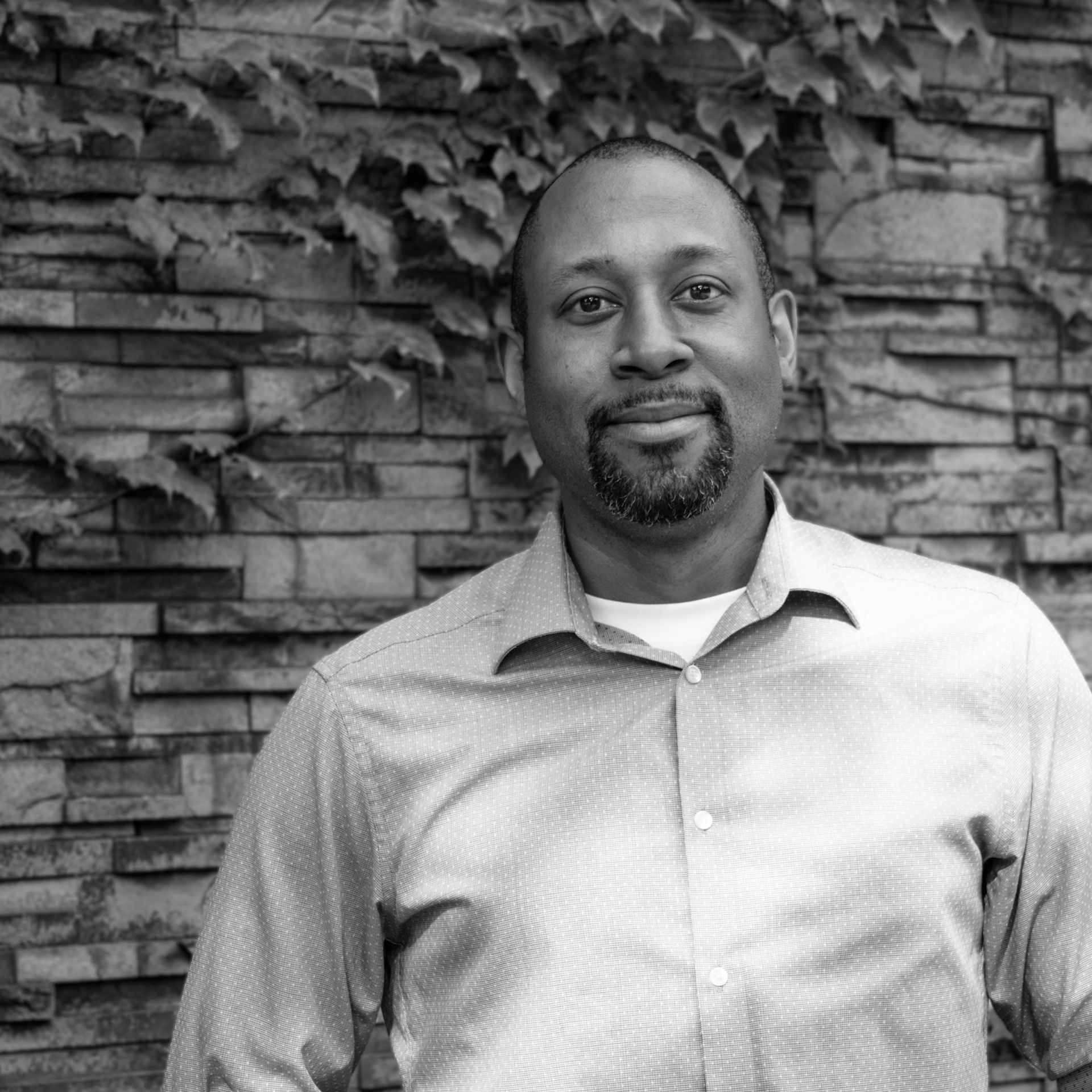 A man with a beard is standing in front of a brick wall in a black and white photo.