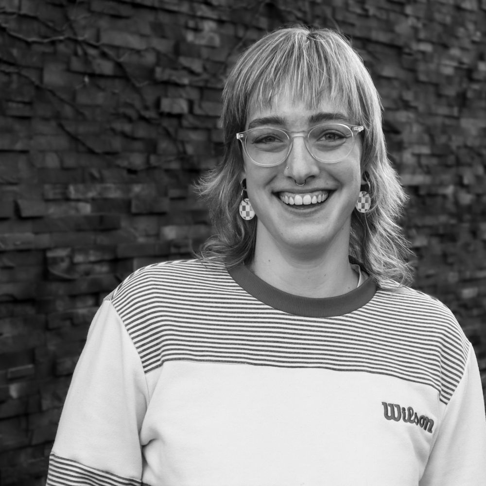 A woman wearing glasses and a wilson shirt is smiling in front of a brick wall.