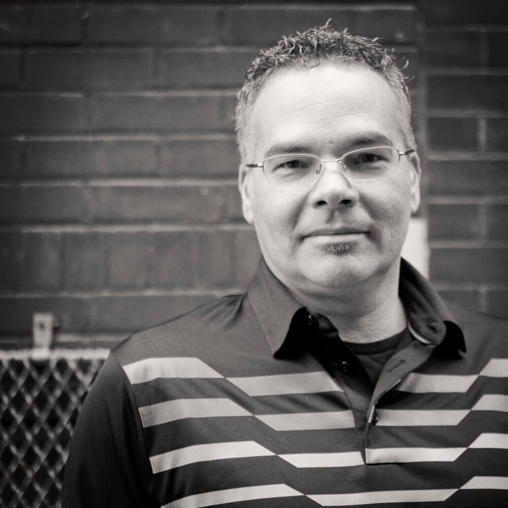 A man wearing glasses and a striped shirt is standing in front of a brick wall