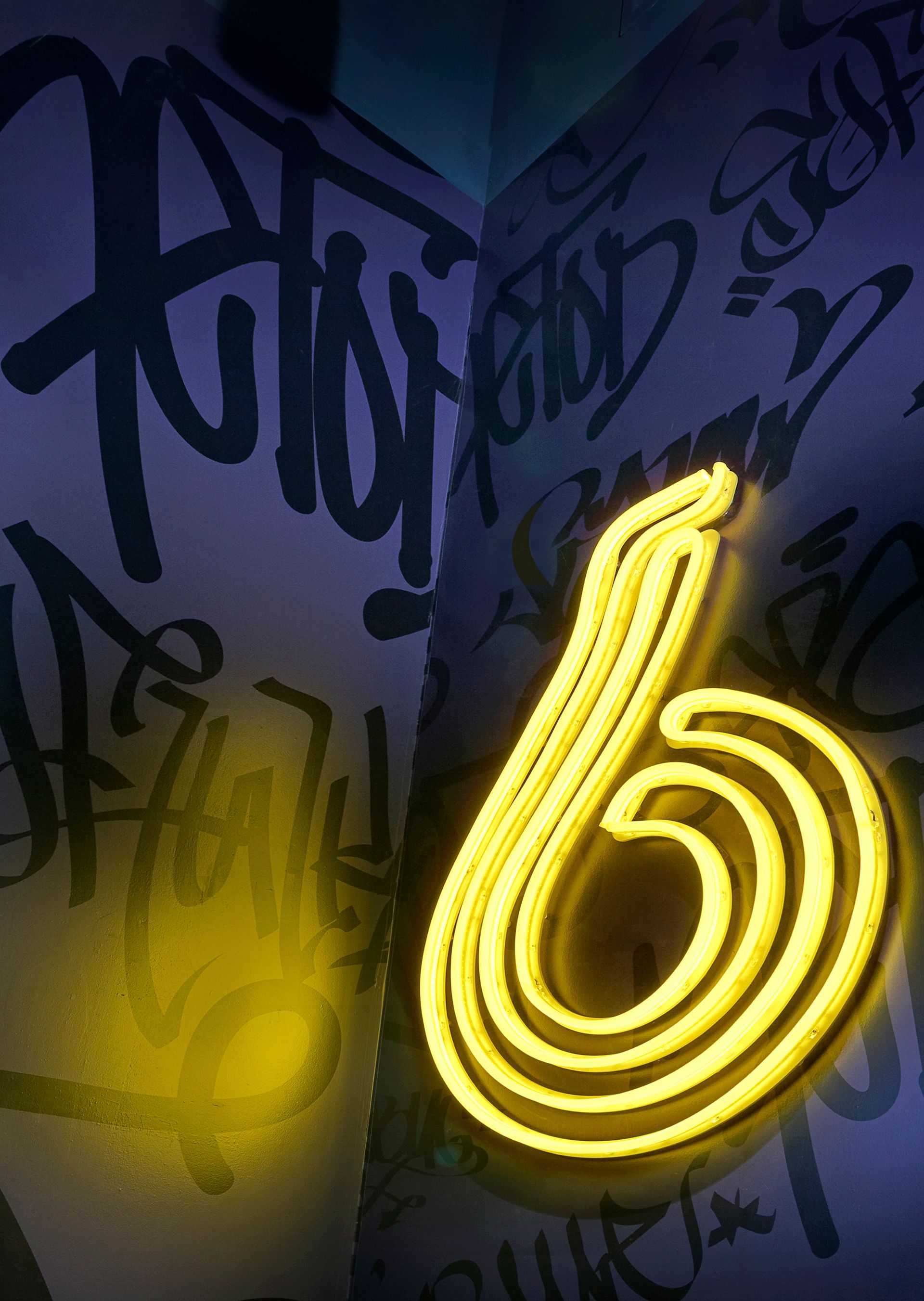 Glowing yellow neon lowercase “6” on a dark purple graffiti-covered wall