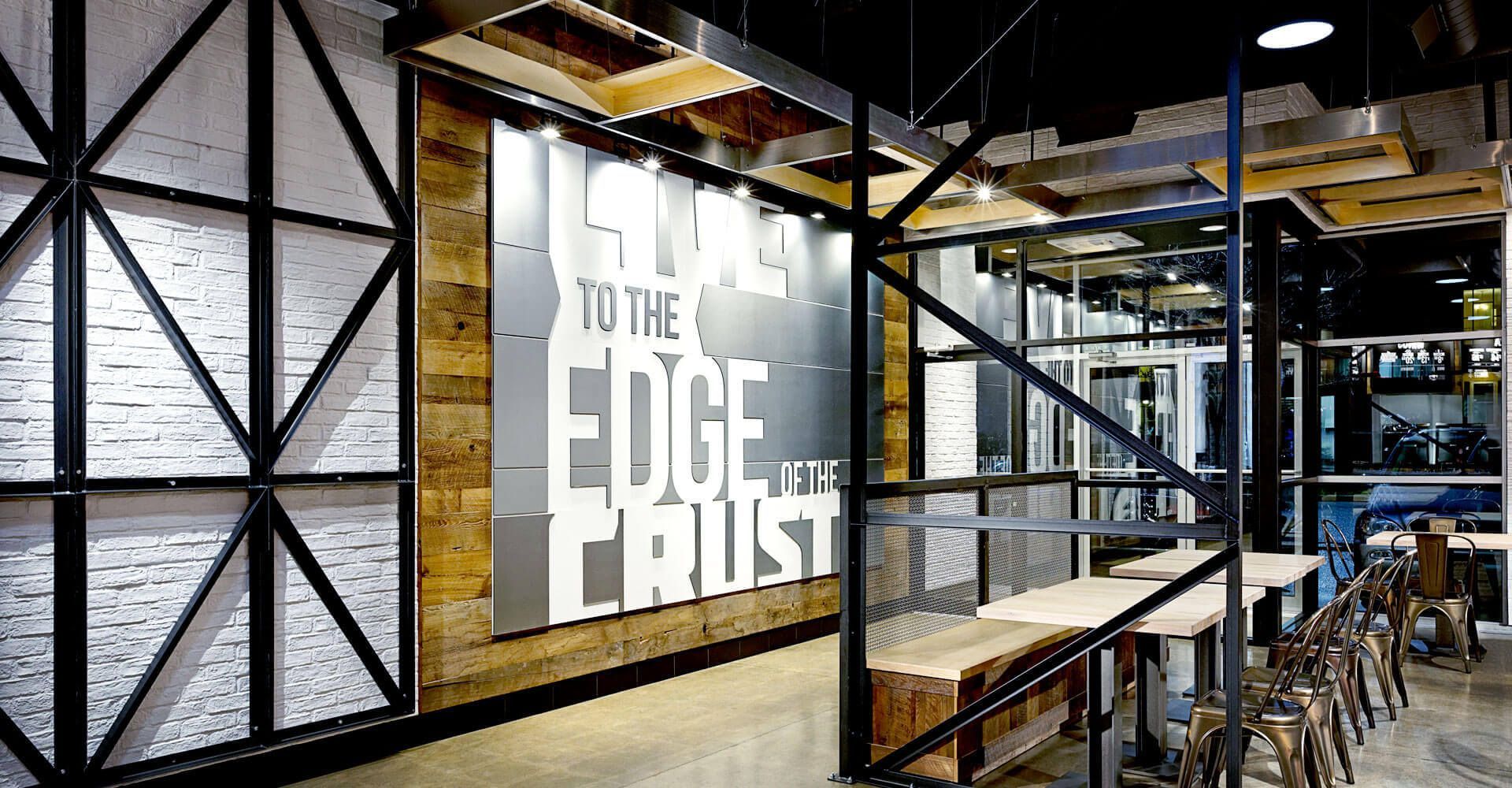 Industrial-style café interior with metal shelves and a large wall sign reading “Live to the Edge of the Crust.”