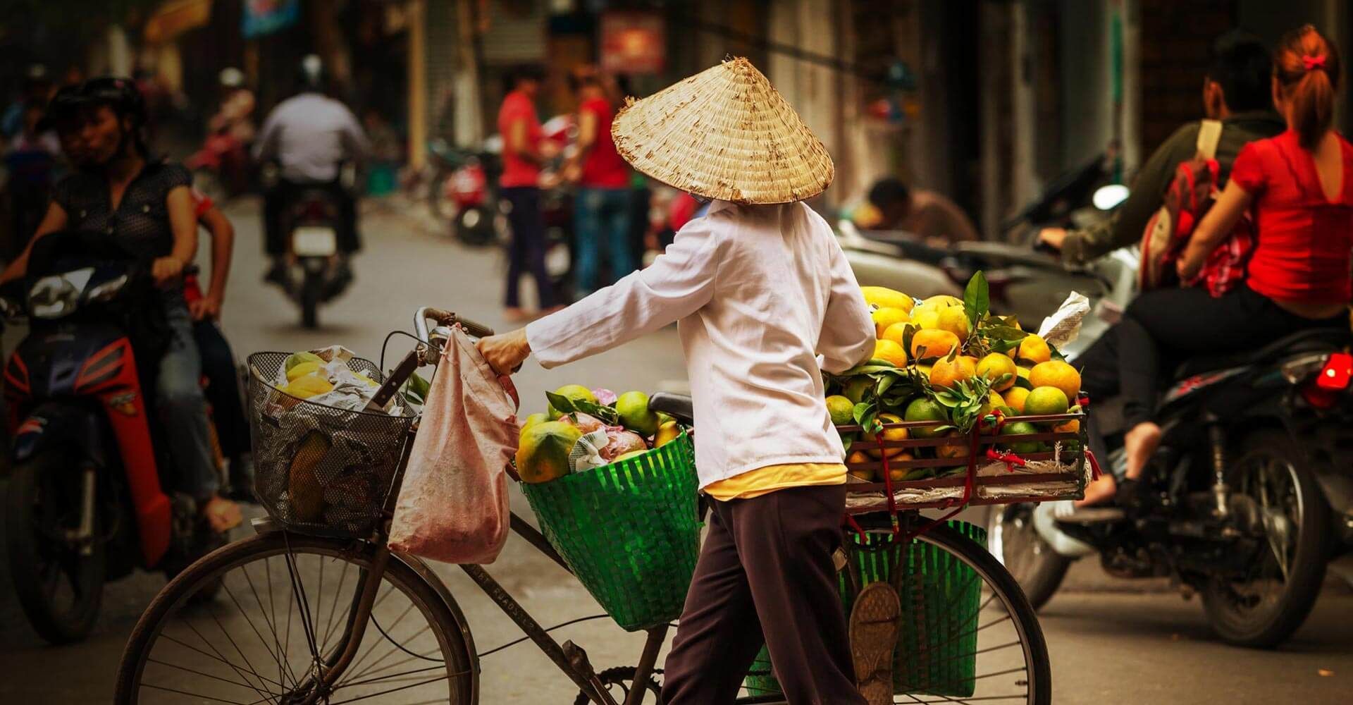 A woman wearing a conical hat is riding a bike down a busy street.