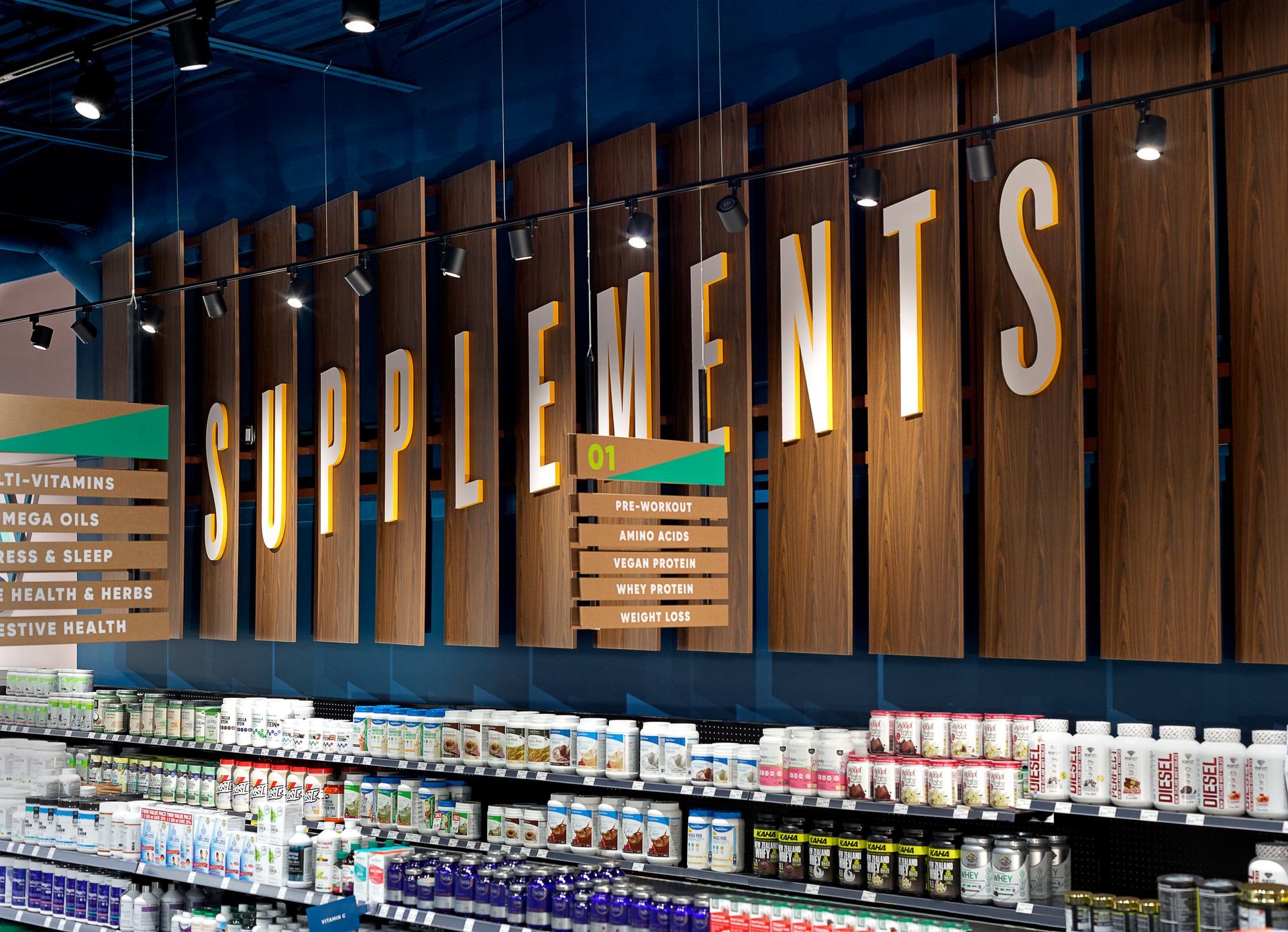 Grocery store supplement aisle with a large “SUPPLEMENTS” sign above stocked shelves.