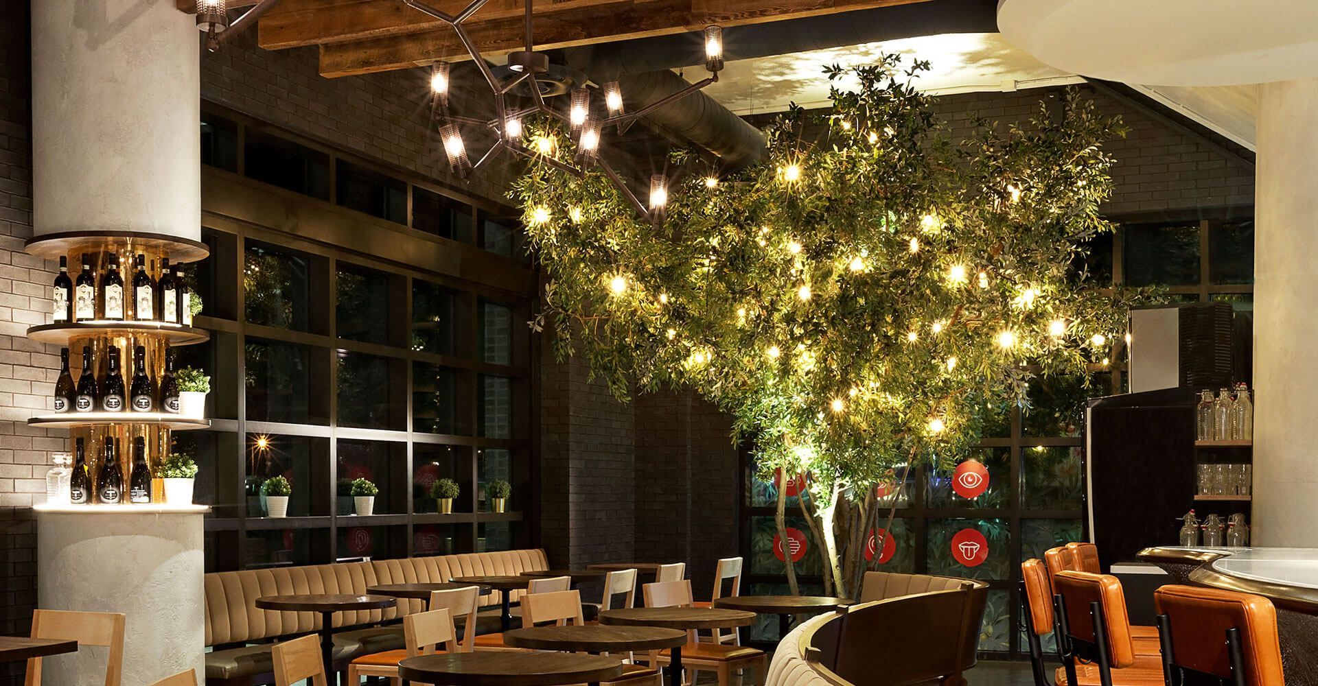 Modern restaurant interior with warm lighting, wooden tables, and a large indoor tree wrapped in fairy lights