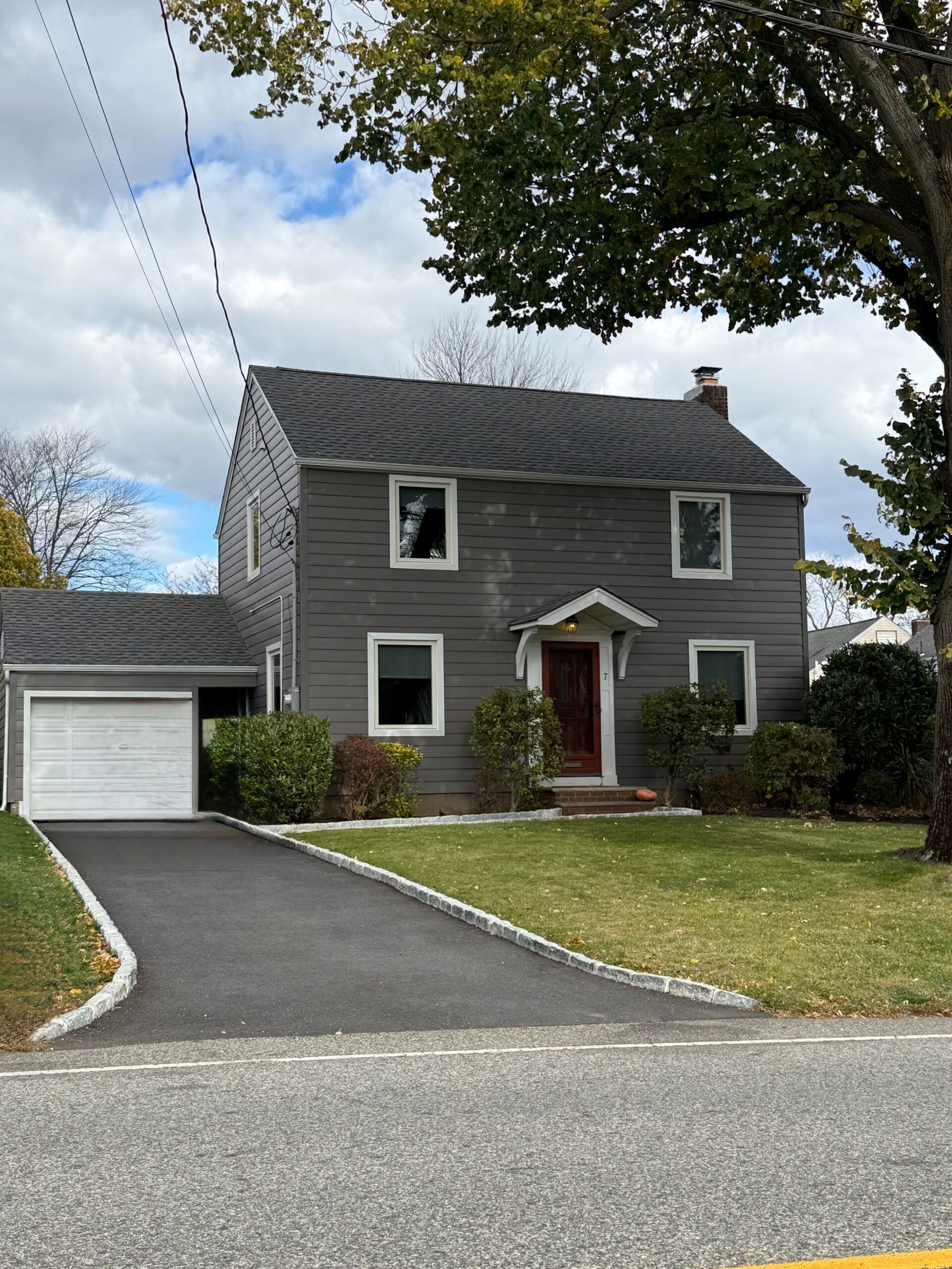 Gray two-story house with a driveway and attached garage. Green grass and trees frame the scene. Cloudy sky above.