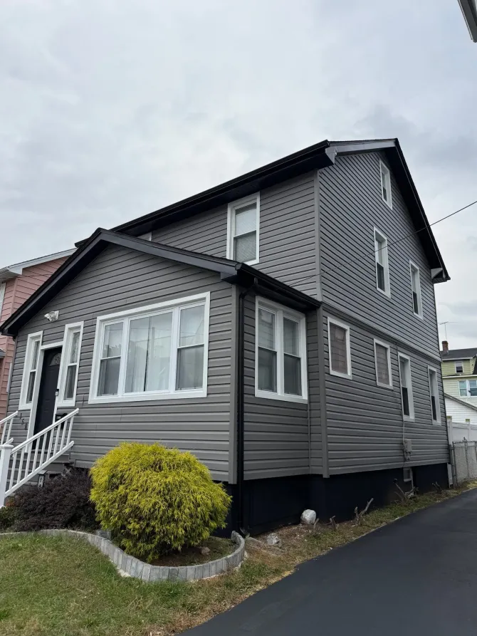 Gray two-story house with white trim and porch beside a paved road under an overcast sky with new siding by American Home Remodeling NJ