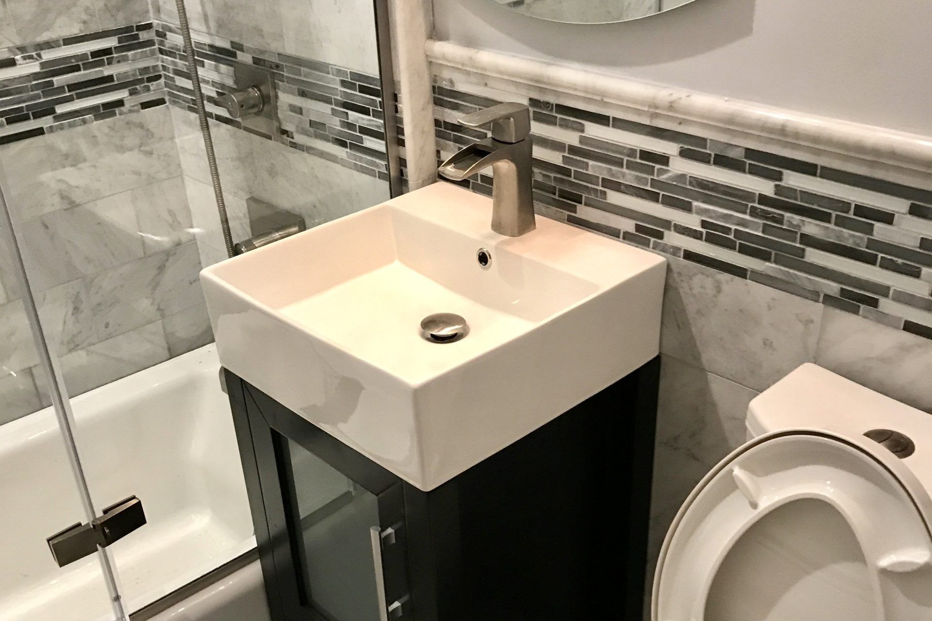 Modern bathroom with glass shower, white square sink on black cabinet, and toilet with mosaic tile backsplash
