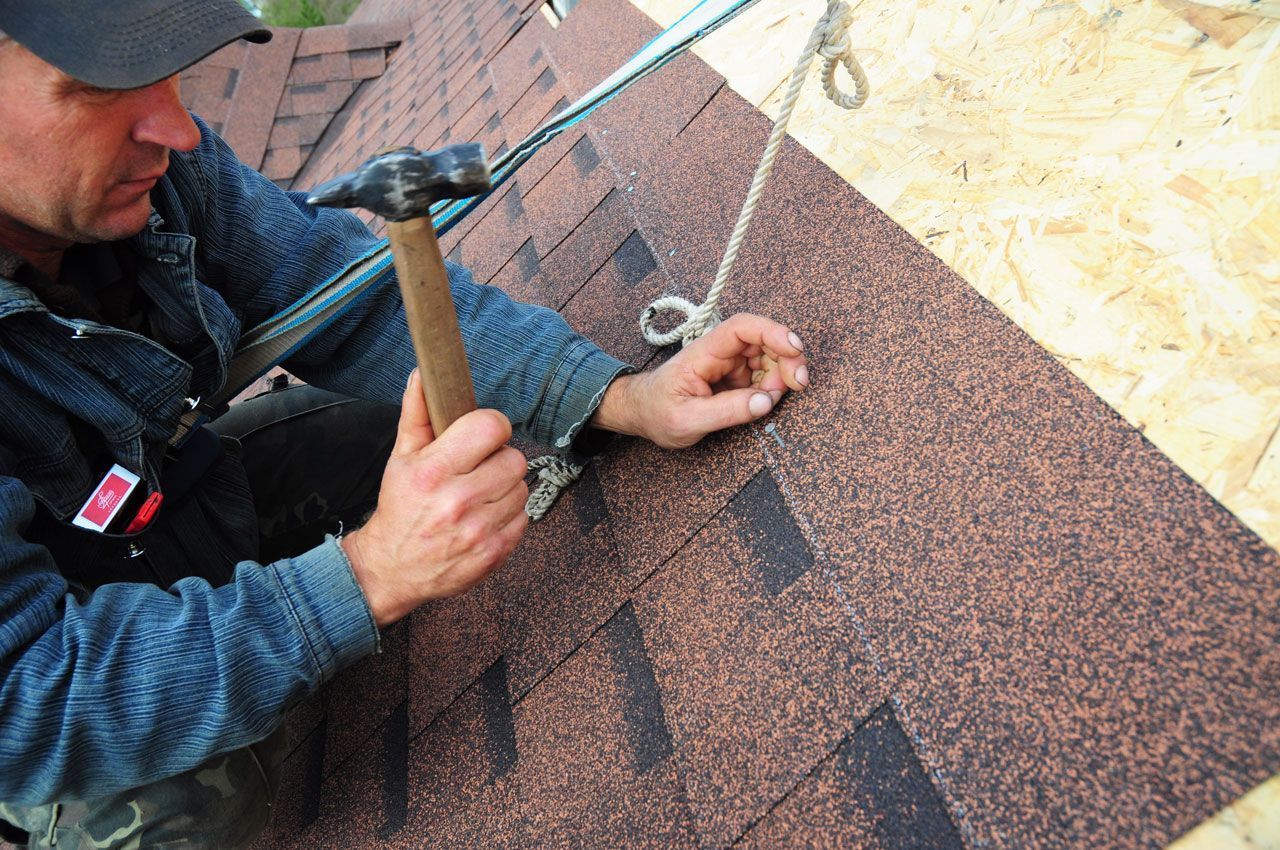 Roofer hammering a shingle. He wears a safety harness on a wooden roof, brown shingles visible.