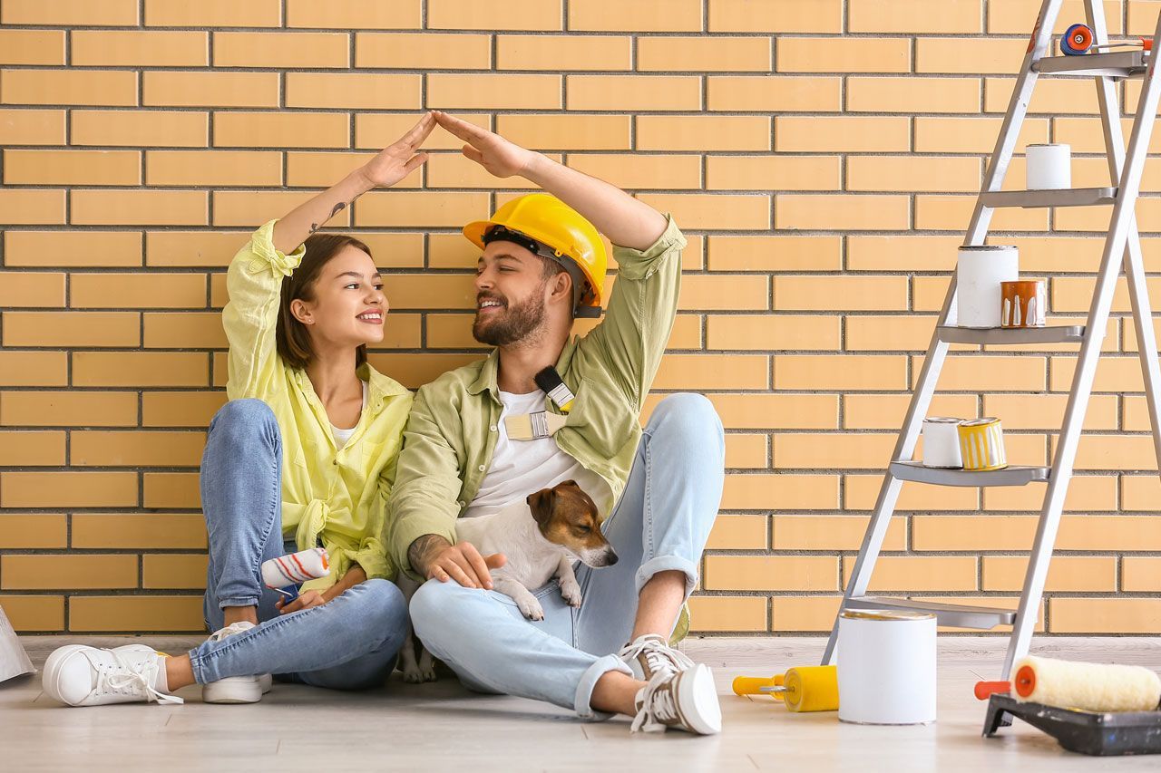 Couple making roof shape with arms, sitting against brick wall with dog, ladder, and paint supplies.