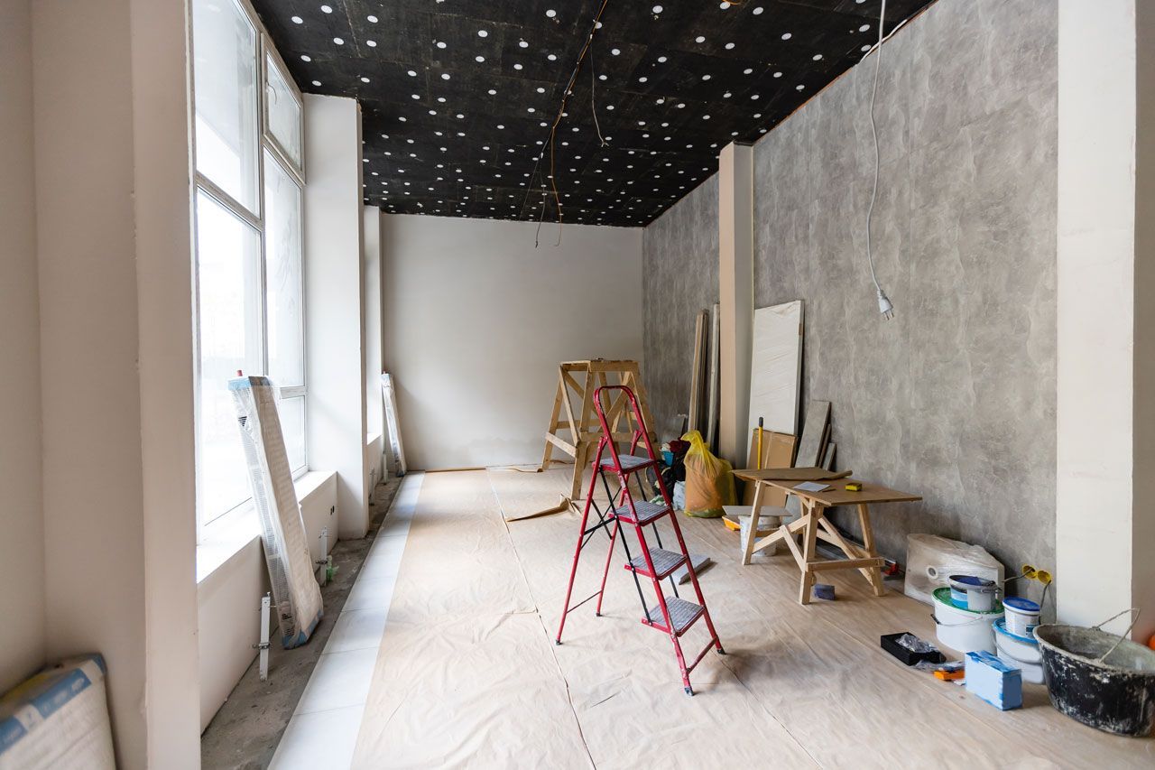 Room under construction with a stepladder, tools, and materials. Large windows and textured walls are visible.